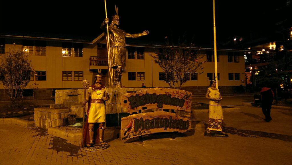 Aguas Calientes: main square at night with a golden statue of an Inca ruler on a stone base, two smaller costumed figures and a Bienvenidos a Machupicchu sign