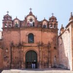 Stone church facade with arched green doors and bell gables on the Plaza de Armas in Cusco, under a clear blue sky
