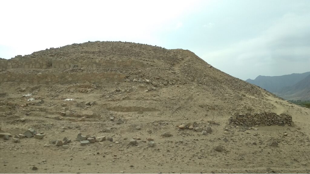 Caral: a large mound of stone and earth on dry desert ground under a pale, hazy sky, with low hills in the distance