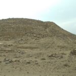 Caral: a large mound of stone and earth on dry desert ground under a pale, hazy sky, with low hills in the distance