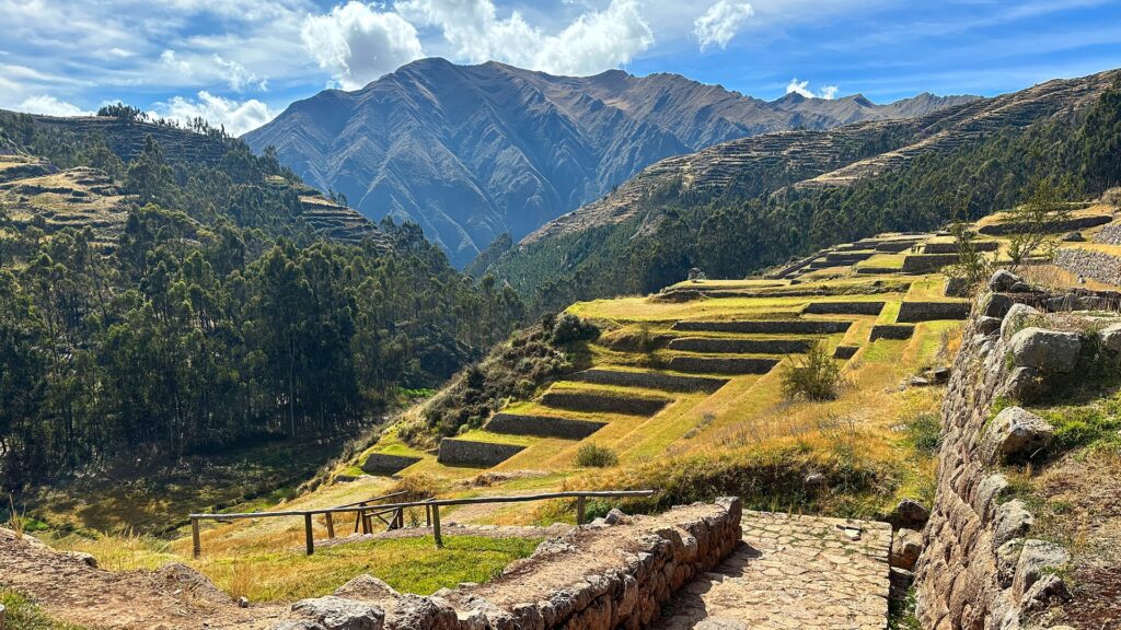 Chinchero: green and golden Inca farming terraces curving down a hillside, a stone wall and stone path in front, and tall mountains under a blue sky