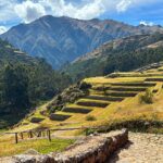 Chinchero: green and golden Inca farming terraces curving down a hillside, a stone wall and stone path in front, and tall mountains under a blue sky
