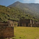 Choquequirao: Inca stone buildings and terrace walls beside a flat green lawn, with a steep forested mountain rising behind under a blue sky