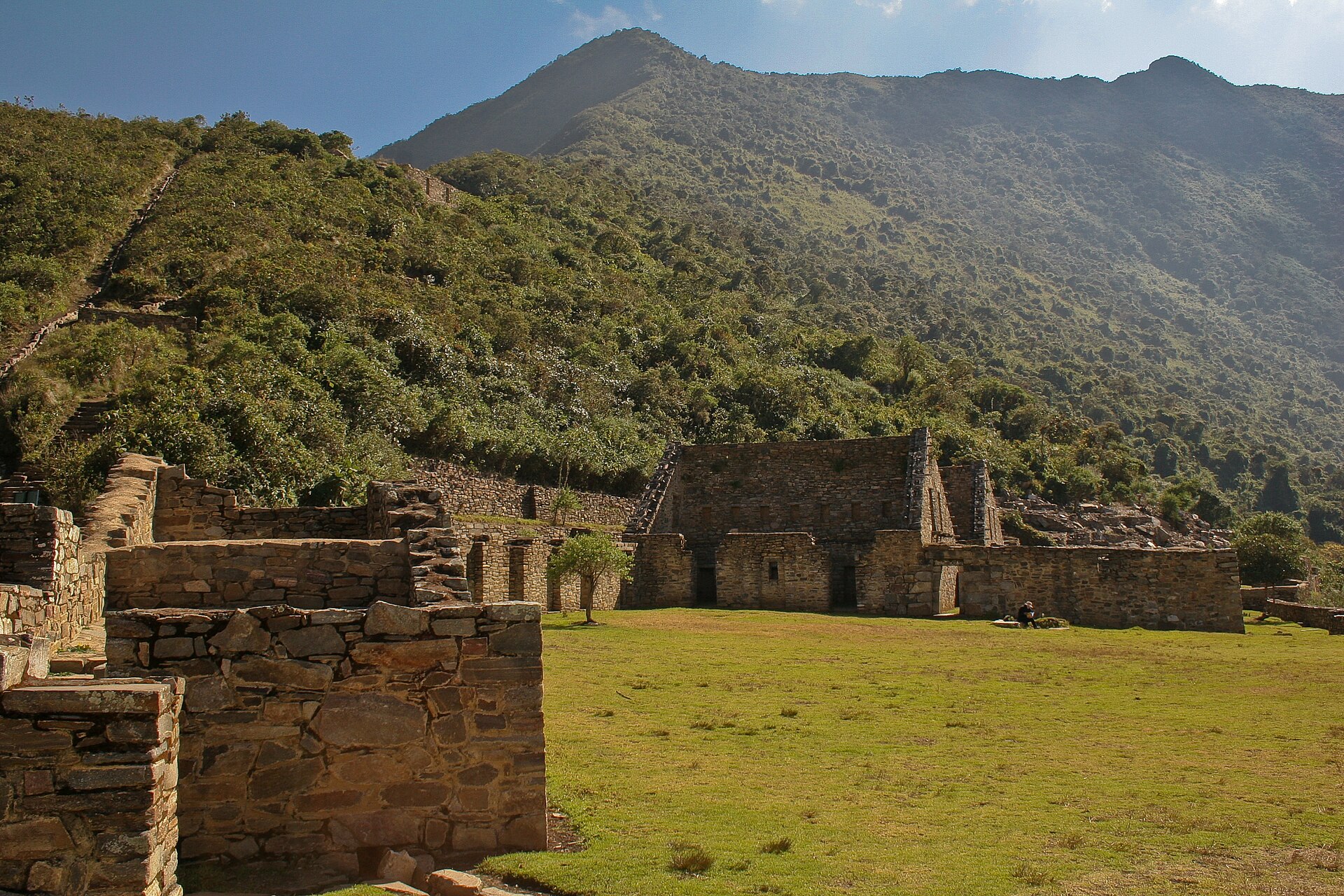 Choquequirao: Inca stone buildings and terrace walls beside a flat green lawn, with a steep forested mountain rising behind under a blue sky
