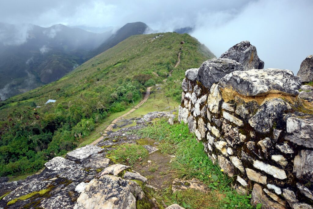 Kuelap: tall stone wall of the Chachapoya citadel on a grassy ridge, with a dirt path and green mountains under low clouds