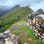 Kuelap: tall stone wall of the Chachapoya citadel on a grassy ridge, with a dirt path and green mountains under low clouds