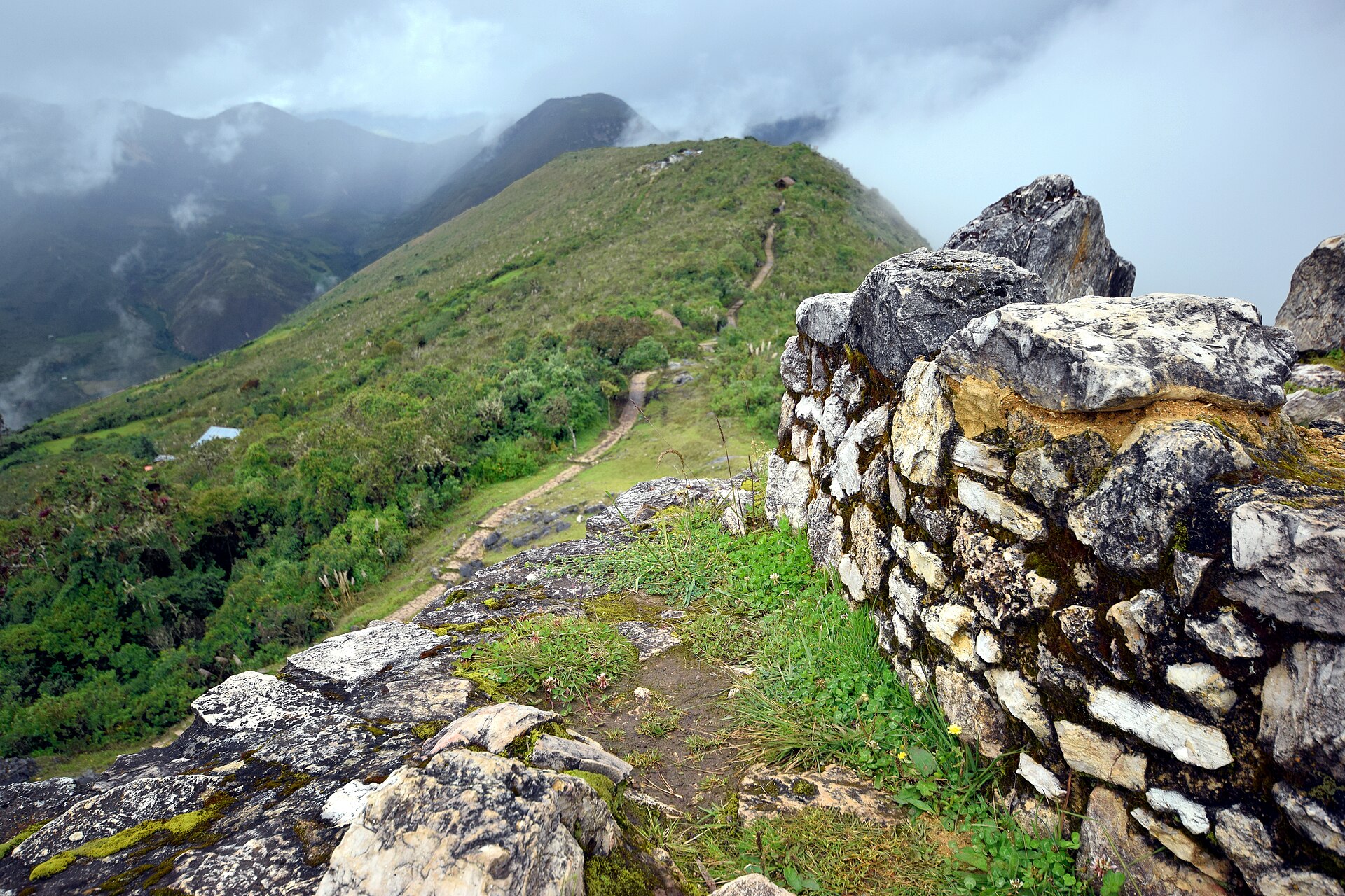 Kuelap: tall stone wall of the Chachapoya citadel on a grassy ridge, with a dirt path and green mountains under low clouds