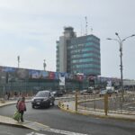 Road and terminal buildings at Jorge Chávez International Airport in Lima, with the control tower and a tall glass office building under an overcast sky