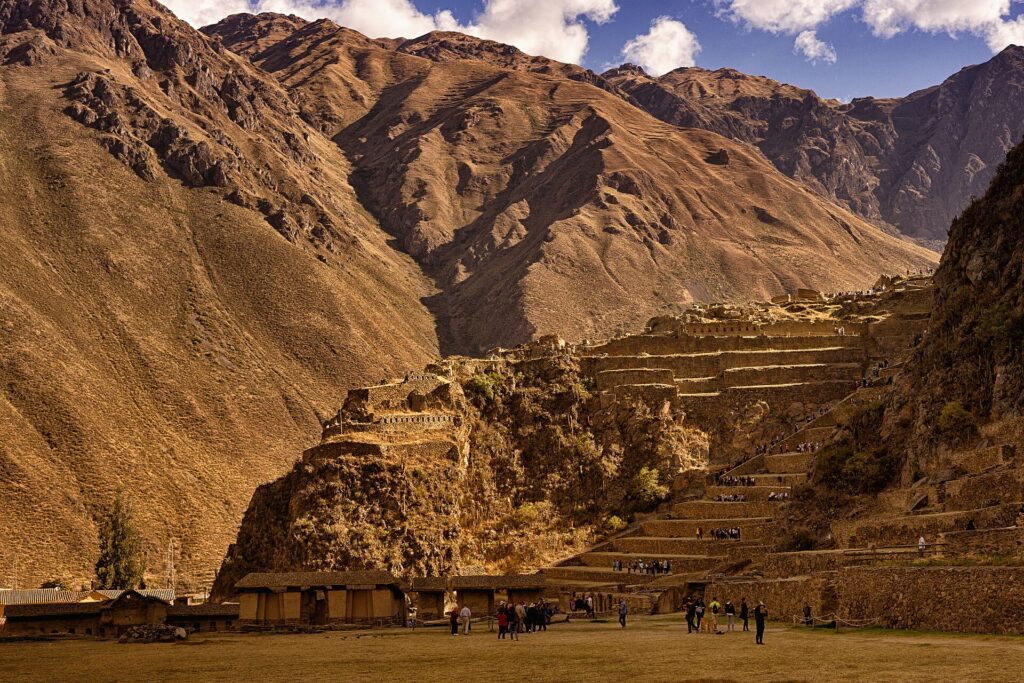 Ollantaytambo's stone Inca terraces climbing a steep, dry hillside, with visitors on the esplanade and brown mountains under a partly cloudy sky