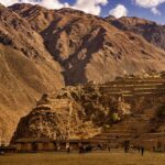 Ollantaytambo's stone Inca terraces climbing a steep, dry hillside, with visitors on the esplanade and brown mountains under a partly cloudy sky