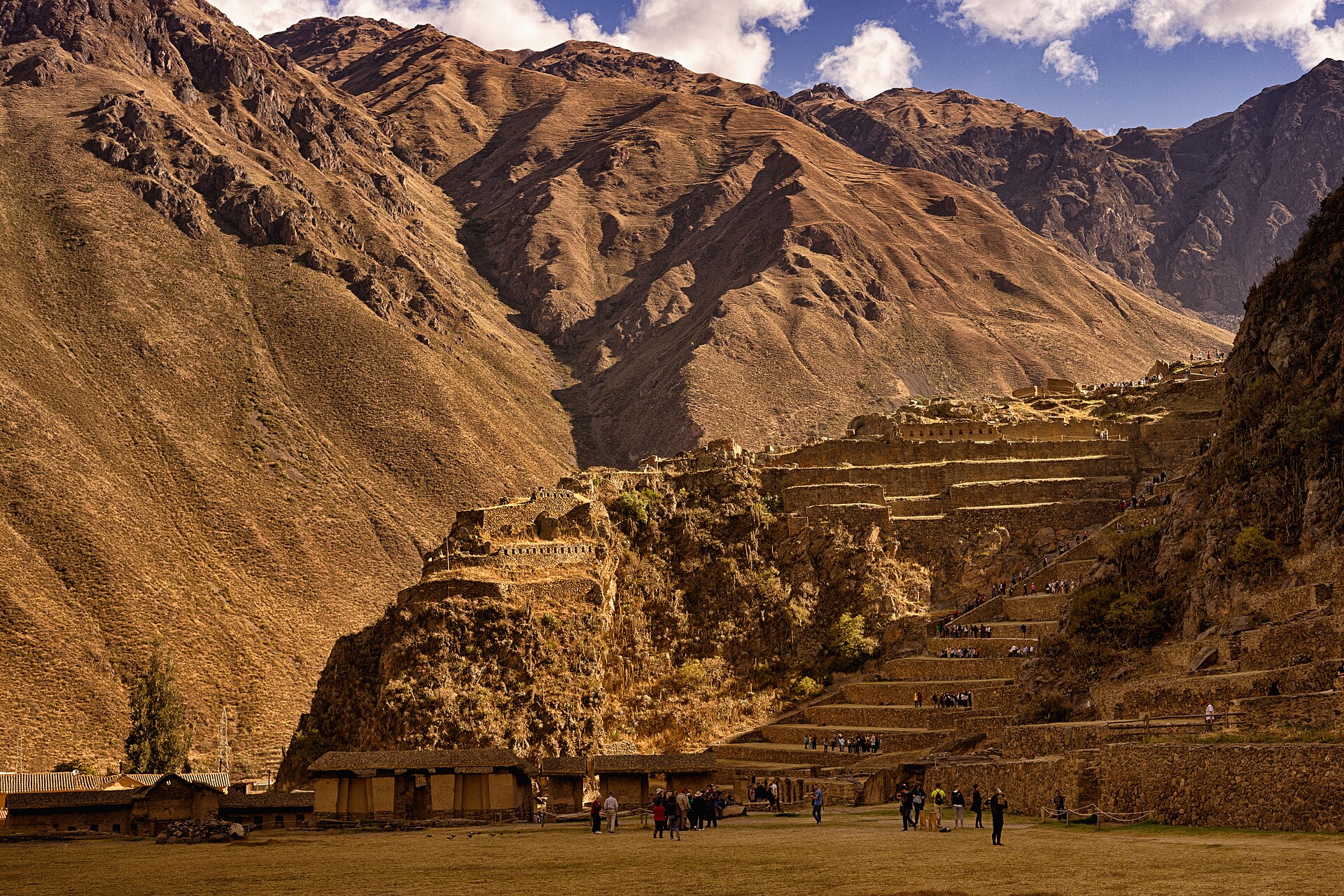 Ollantaytambo's stone Inca terraces climbing a steep, dry hillside, with visitors on the esplanade and brown mountains under a partly cloudy sky