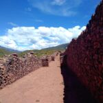 Pikillaqta: a long dirt street between high walls of rough reddish stone, with green hills and white clouds in a blue sky behind