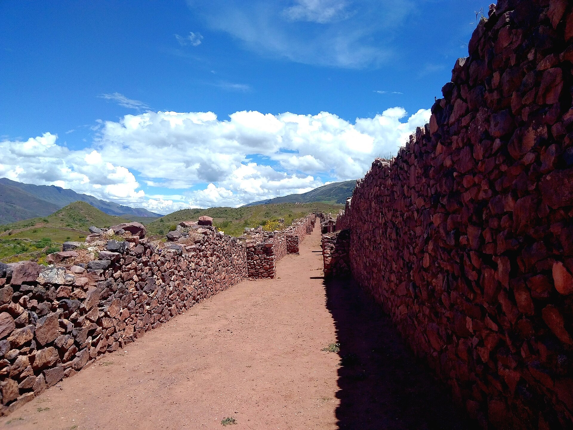 Pikillaqta: a long dirt street between high walls of rough reddish stone, with green hills and white clouds in a blue sky behind