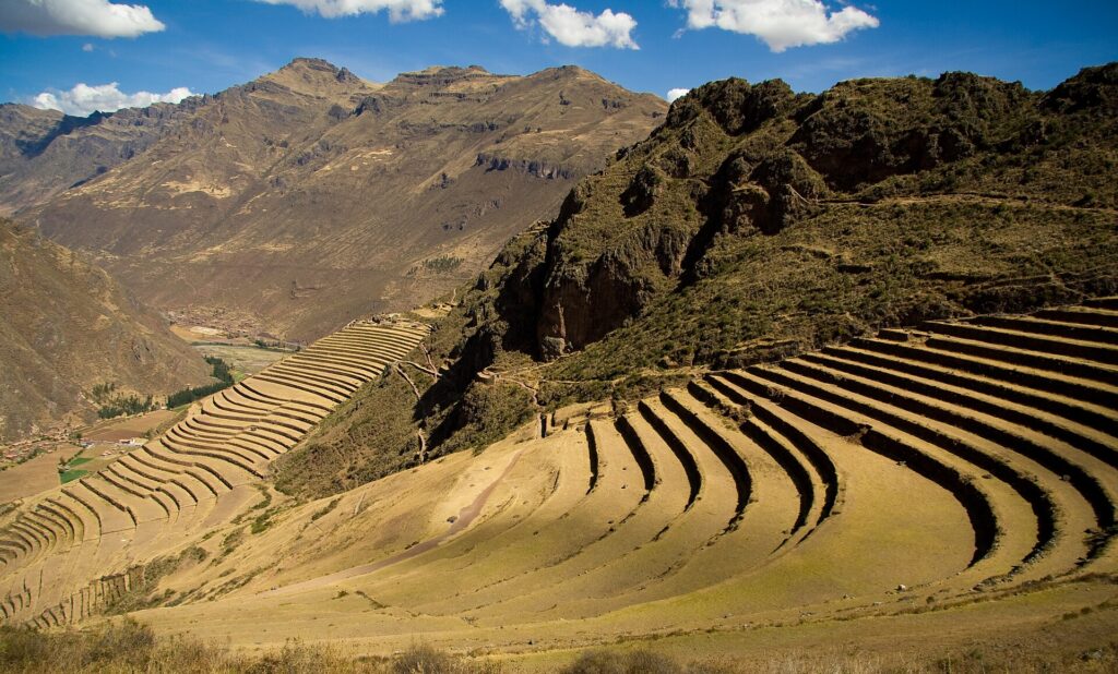 Pisac: curved Inca farming terraces climbing a dry hillside, with a rocky ridge on the right and brown mountains under a blue sky with white clouds