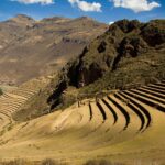 Pisac: curved Inca farming terraces climbing a dry hillside, with a rocky ridge on the right and brown mountains under a blue sky with white clouds