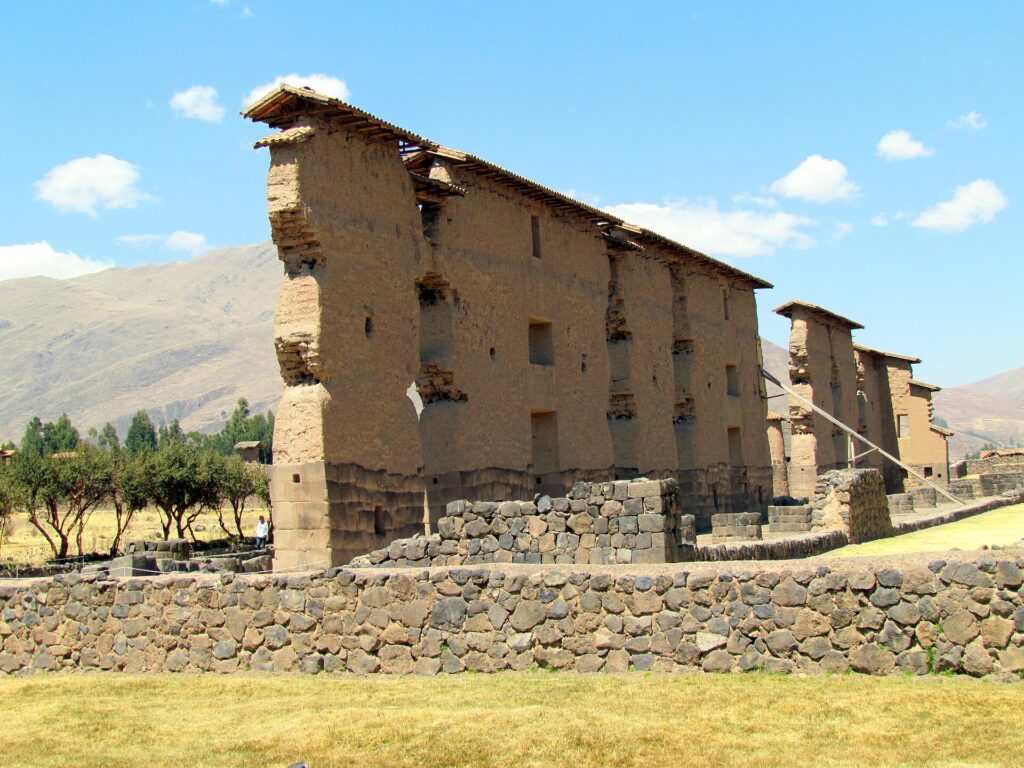 Raqchi: the tall Temple of Viracocha wall, with a stone base, an adobe upper part and a small roof, behind a low stone wall and dry grass under a blue sky