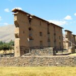 Raqchi: the tall Temple of Viracocha wall, with a stone base, an adobe upper part and a small roof, behind a low stone wall and dry grass under a blue sky