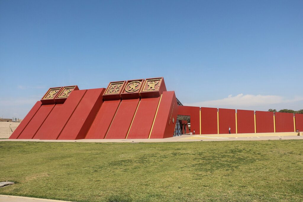 Royal Tombs of Sipan Museum: red pyramid-shaped building with gold Moche reliefs on top, a long red wall and a green lawn under a blue sky