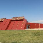 Royal Tombs of Sipan Museum: red pyramid-shaped building with gold Moche reliefs on top, a long red wall and a green lawn under a blue sky