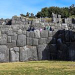 Sacsayhuaman: tiers of huge grey Inca stone walls above a green lawn, with visitors walking on an upper terrace and trees under a blue sky