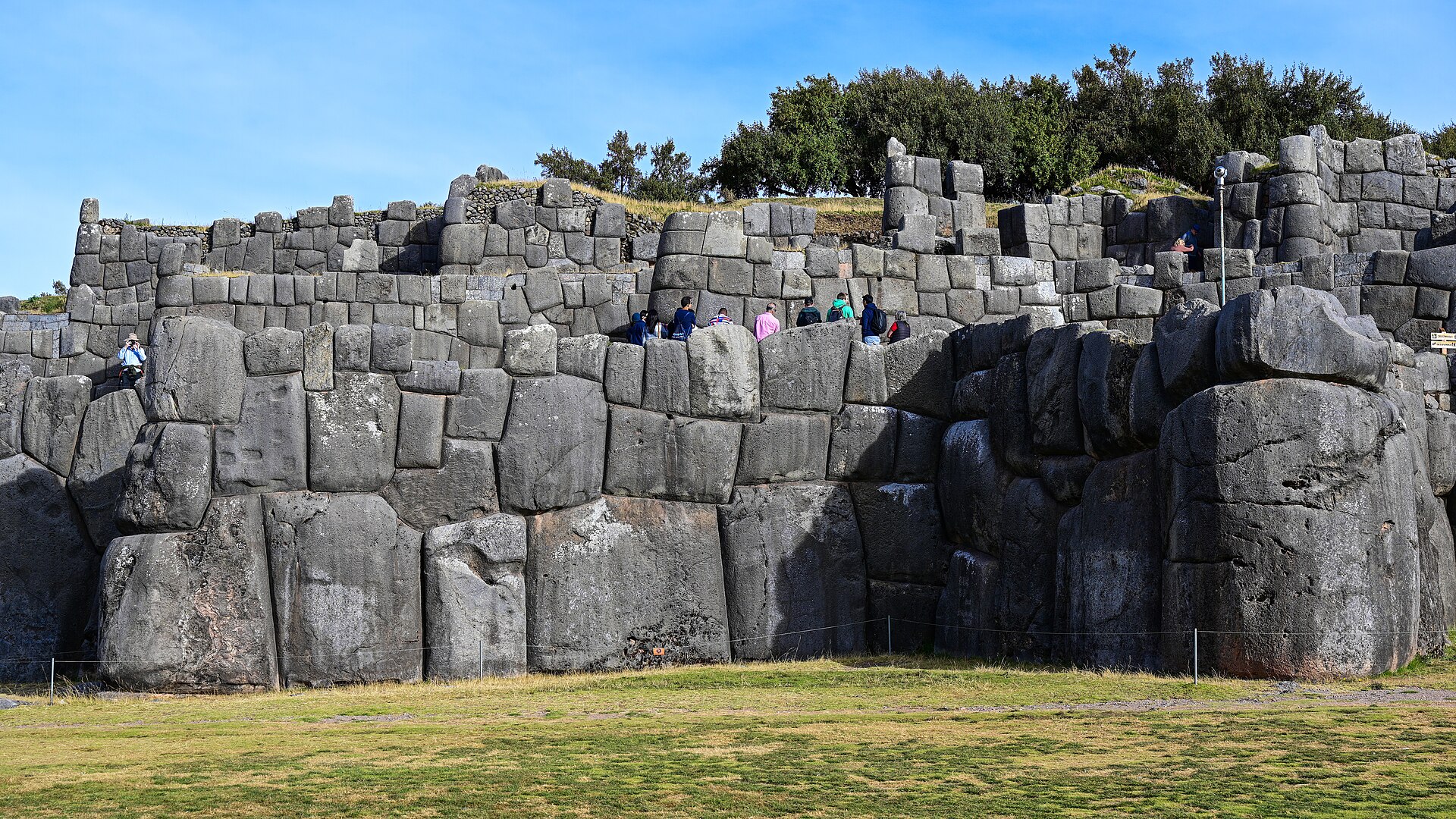 Sacsayhuaman: tiers of huge grey Inca stone walls above a green lawn, with visitors walking on an upper terrace and trees under a blue sky
