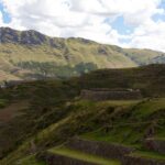 Tipon: Inca stone terrace walls and a round stone structure on a green hillside, with a long grassy mountain ridge and white clouds behind
