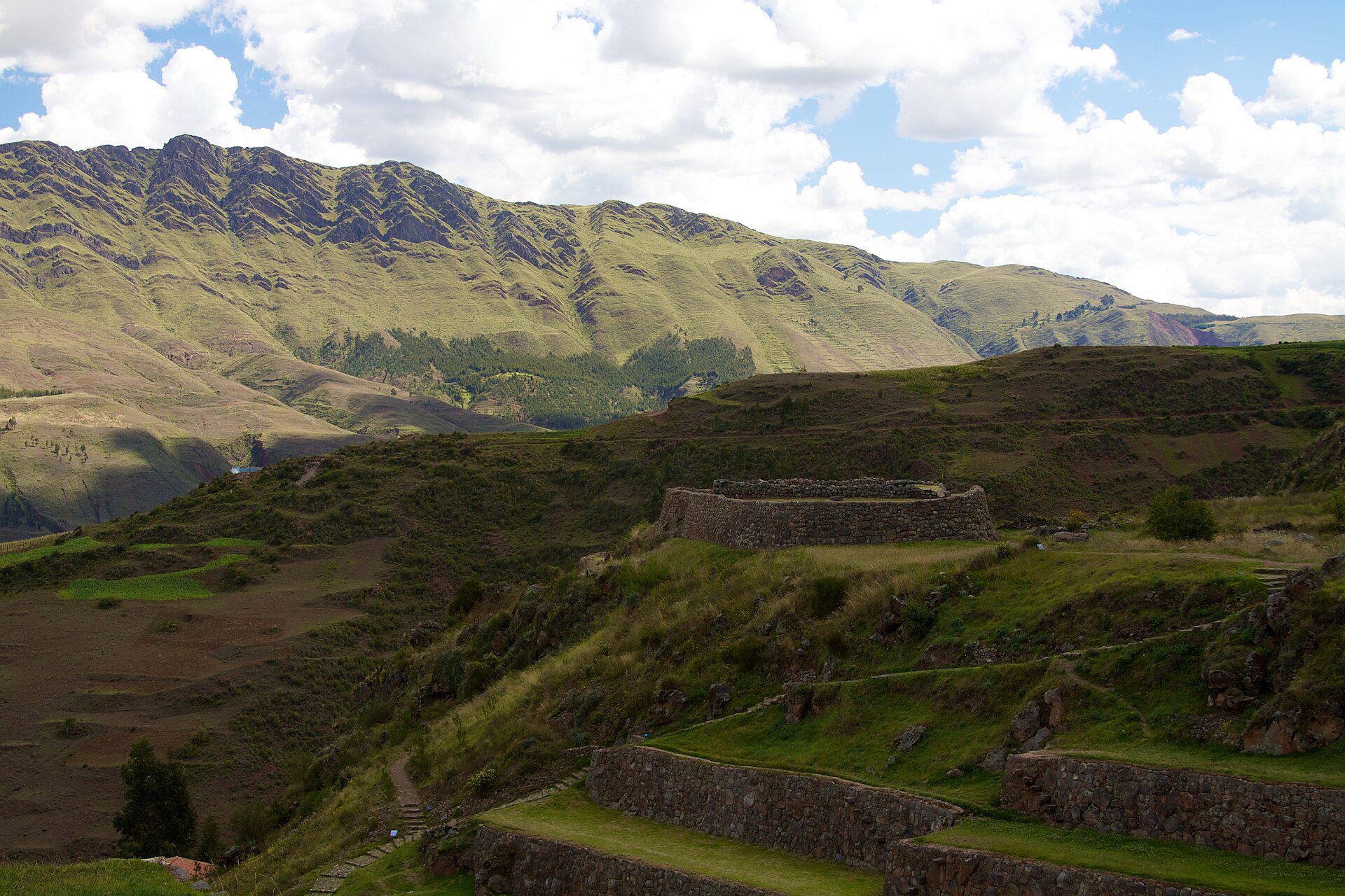 Tipon: Inca stone terrace walls and a round stone structure on a green hillside, with a long grassy mountain ridge and white clouds behind
