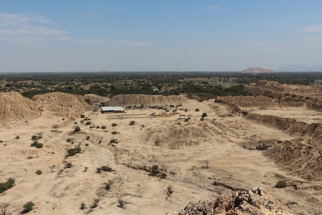 Túcume: eroded adobe pyramids and mounds on a flat sandy plain with scattered shrubs and a small building, seen from above under a pale blue sky