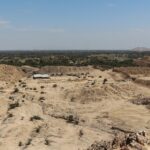 Túcume: eroded adobe pyramids and mounds on a flat sandy plain with scattered shrubs and a small building, seen from above under a pale blue sky