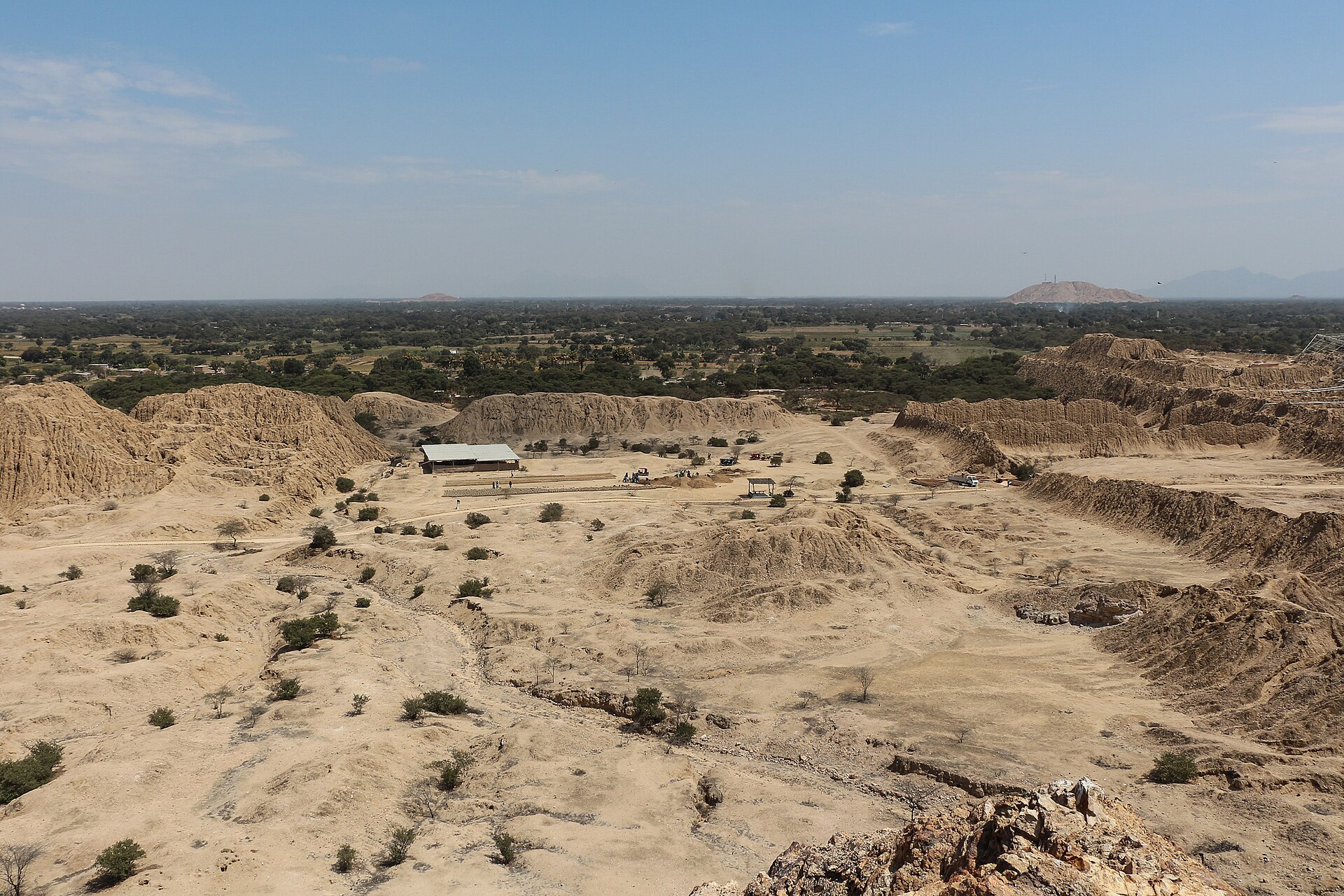 Túcume: eroded adobe pyramids and mounds on a flat sandy plain with scattered shrubs and a small building, seen from above under a pale blue sky