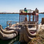 Reed boats and a wooden tower with two people on a Uros floating island, with the blue water of Lake Titicaca behind