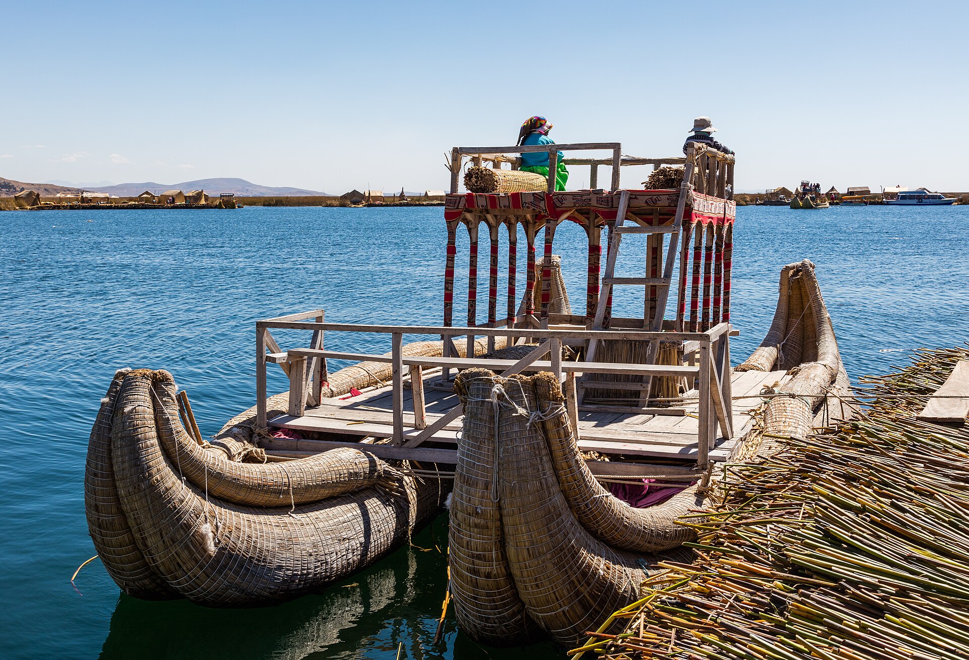 Reed boats and a wooden tower with two people on a Uros floating island, with the blue water of Lake Titicaca behind